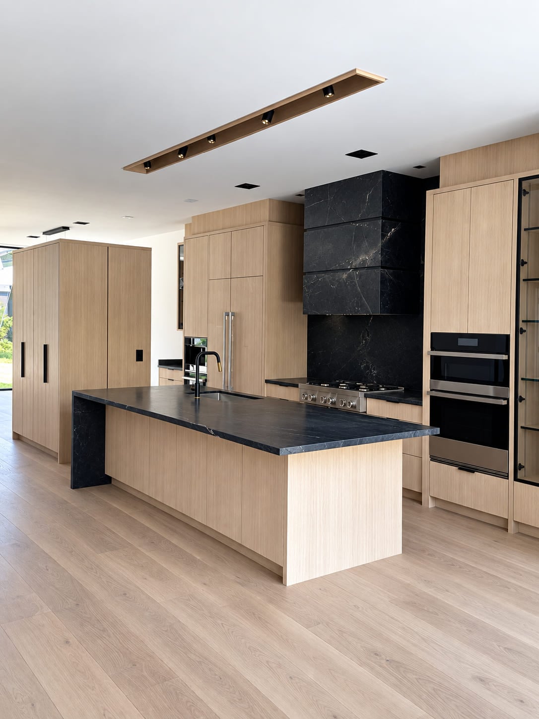 Kitchen island in dark veined stone with the top turning down at the end and running to the floor as a waterfall panel, in pale oak cabinetry